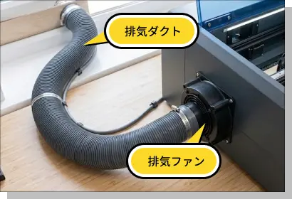 レーザーカッターの排気システム。マシン背面の排気ファンから太いグレーの排気ダクトが伸び、屋外へ煙を排出する接続の様子。