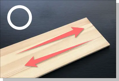 正しいサンディングの方向を示す解説写真。木材の繊維（木目）の流れに沿って、赤い矢印が平行に往復している様子。