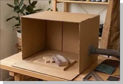 段ボールで作った簡易的なサンディングブース。右側に掃除機のダクトが接続されており、内部には研磨用の木片とサンディングスポンジが置かれている様子。